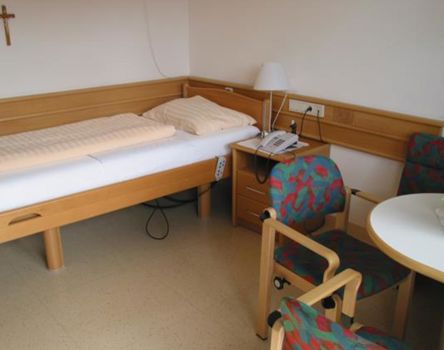 Die Einrichtung eines Zimmers: ein höhenverstellbares Einzelbett, ein Nachtschränkchen mit Telefonanlage, eine Sitzgruppe und ein Kruzifix an der Wand neben dem Bett