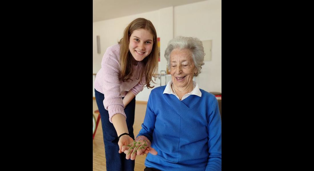 Eine alte Frau zeigt stolz ihre bemalte Hand; eine junge Frau hilft ihr dabei