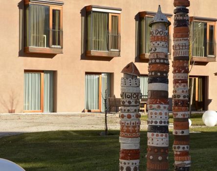 La facciata del centro residenziale di cura Brunico con ampie finestre e balconi; in primo piano tre colonne artistiche in terracotta colorata su un prato verde, accanto a sfere decorative bianche