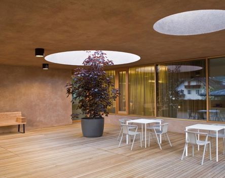 Moderne überdachte Terrasse mit Holzfußboden, weißen Tischen und Stühlen, einer Holzbank und einem Baum im Topf, der durch eine runde Öffnung in der Decke wächst; große Fenster spiegeln die Außenansicht wider.